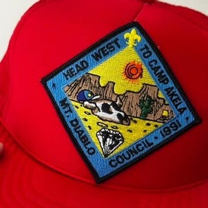 Könnte beinhalten: Rote Trucker-Mütze mit einem quadratischen Aufnäher mit einer Wüstenlandschaft, einem Diamanten und dem Text "HEAD WEST", "TO CAMP AKELA", "MT. DIABLO" und "COUNCIL 1991". Der Aufnäher hat einen blauen Rand und gelbe und braune Details.