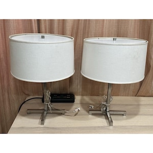May include: A pair of table lamps featuring cylindrical white fabric shades and brushed metal bases with a cross-shaped stand. Each lamp has a central vertical post supporting the shade and a clear power cord extending from the base.