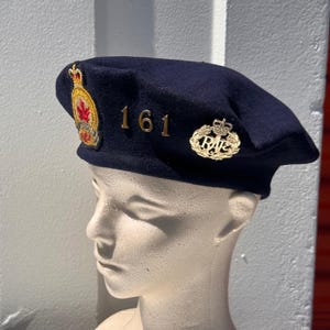 Op de afbeelding: Een donkerblauwe wollen baret met een goud en rood geborduurd embleem aan de linkerkant, de gouden metalen cijfers 161 in het midden en een gouden metalen RAF-badge aan de rechterkant. De baret wordt getoond op een neutraal gekleurd etalagehoofd.