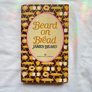May include: A paperback cookbook titled Beard on Bread by James Beard. The cover features a dark brown background with a repeating gold lattice pattern containing various bread illustrations. A large central oval contains the title and author text.