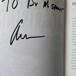 May include: An open book page with handwritten text in black ink. The text reads "TO Dr M Spurr" above a signature. The page is from a book with printed text visible on the right side.
