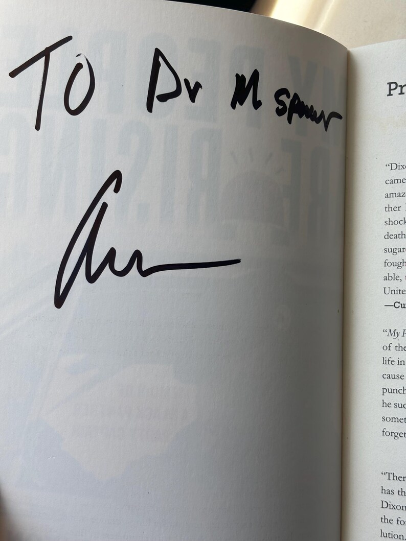 May include: An open book page with handwritten text in black ink. The text reads "TO Dr M Spurr" above a signature. The page is from a book with printed text visible on the right side.