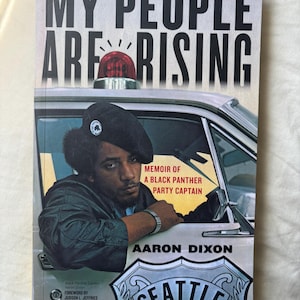 May include: A book titled "MY PEOPLE ARE RISING" with a photo of a Black Panther Party Captain. The cover features a police car and the text "MEMOIR OF A BLACK PANTHER PARTY CAPTAIN" by Aaron Dixon. The city of Seattle is also visible.