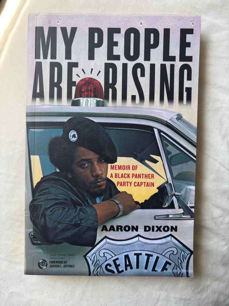 May include: A book titled "MY PEOPLE ARE RISING" with a photo of a Black Panther Party Captain. The cover features a police car and the text "MEMOIR OF A BLACK PANTHER PARTY CAPTAIN" by Aaron Dixon. The city of Seattle is also visible.