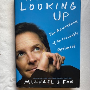 May include: A book cover with a blue background and the title "ALWAYS LOOKING UP" in white. The book features a photo of Michael J. Fox and the subtitle "The Adventures of an Incurable Optimist". The author's name is also displayed.