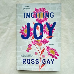 May include: A book titled Inciting Joy by Ross Gay with a white cover featuring a light gray brick pattern. The title is printed in large blue letters, surrounded by illustrations of bright pink flowers with yellow centers and green stems.