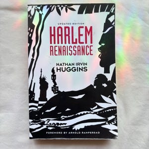May include: A paperback book titled Harlem Renaissance by Nathan Irvin Huggins, updated edition. The cover features a black and white graphic illustration of a reclining figure, a crocodile, and the Statue of Liberty, with red text for the title.