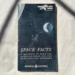 May include: A dark blue booklet titled SPACE FACTS: A HANDBOOK OF BASIC AND ADVANCED SPACE DATA FOR SCIENTISTS AND ENGINEERS. The cover features a graphic of a moon, stars, and the text MISSILE AND SPACE VEHICLE DEPARTMENT GENERAL ELECTRIC.