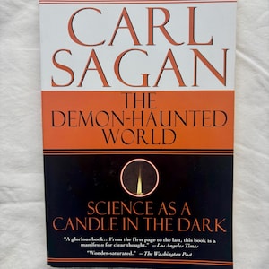 May include: A paperback book titled The Demon-Haunted World: Science as a Candle in the Dark by Carl Sagan. The cover features a white top section, an orange middle band, and a black bottom section with a graphic of a single lit candle in a circle.