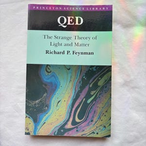 May include: A paperback book titled QED The Strange Theory of Light and Matter by Richard P. Feynman from the Princeton Science Library. The cover features a black top section with white text and a lower section with a colorful oil-slick pattern.