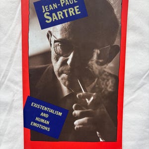 May include: A book cover featuring a black and white portrait of Jean-Paul Sartre, wearing glasses and holding a pipe. The title "Jean-Paul Sartre" is in white text on a blue rectangle. The book's title "Existentialism and Human Emotions" is also on a blue rectangle.