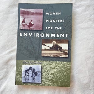 May include: A book titled Women Pioneers for the Environment by Mary Joy Breton. The dark green cover features a large leaf outline and three sepia-toned photographs: two people wading in water, a person kayaking, and two people working in a marsh.