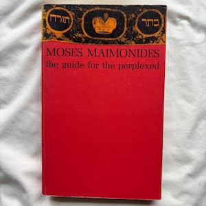 May include: A book titled The Guide for the Perplexed by Moses Maimonides. The cover is bright red with a black top border featuring an ornate gold crown graphic flanked by two circular emblems containing Hebrew text in a decorative style.
