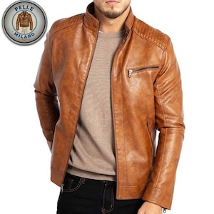 Könnte beinhalten: Eine braune Lederjacke mit Stehkragen und Reißverschluss. Die Jacke hat eine Steppung an den Schultern und eine kleine Brusttasche mit Reißverschluss. Die Jacke wird über einem hellbraunen Pullover getragen. Das Pelle Milano Logo ist sichtbar.