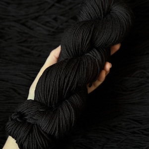 Può includere: Una singola matassa di filato nero tinta unita, attorcigliata strettamente. Il filato presenta una struttura a più capi liscia e coerente con una leggera lucentezza, mostrando un colore nero profondo e uniforme lungo tutta la fibra.