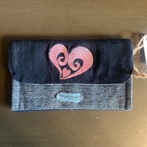 Op de afbeelding: Een denim portemonnee met een roze geborduurd hartontwerp. De portemonnee heeft een klepsluiting en een klein label met de tekst "Hand made with love". De portemonnee is gemaakt van donker- en lichtblauwe denim.