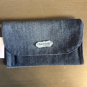 Op de afbeelding: Een denim portemonnee met een klepsluiting. De portemonnee is gemaakt van blauwe spijkerstof en heeft een klein, ovaal label met de tekst "Hand made with Love". De portemonnee is gesloten en ligt op een houten oppervlak.