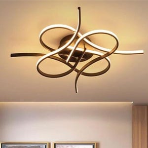 Puede incluir: Lámpara de techo moderna con un diseño abstracto único. La lámpara presenta una serie de brazos curvos y en bucle con iluminación LED integrada. Está montada en un techo de color neutro, iluminando una sala de estar con un sofá de cuero marrón y obras de arte.