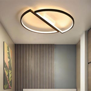 May include: Contemporary ceiling light with a black frame and a white, circular light. The design features a rectangular central light and a geometric, intersecting frame. The light is mounted on a white ceiling, illuminating the room.