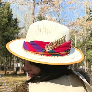 May include: Cream-colored fedora hat with a gold-colored brim and a decorative band. The band features a red, purple, and teal pattern, accented with a gold-colored leaf-shaped embellishment. The hat is a stylish accessory.