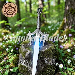 May include: A decorative sword with a silver handle and blade, displayed on a moss-covered rock. The blade has a blue glow. The handle features detailed carvings. The background is a blurred forest. The text "Smoked Blades" is visible.