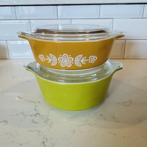 Puede incluir: Dos fuentes de horno redondas de vidrio apiladas con tapas. La fuente superior es de color amarillo mostaza con un patrón floral y de mariposas blanco. La inferior es de color verde lima sólido. Ambas tienen asas laterales y tapas de vidrio.