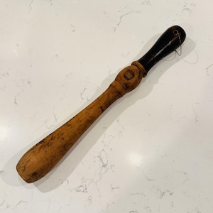 Könnte beinhalten: Ein Holzwerkzeug mit einem langen, hellbraunen, zylindrischen Körper und einem dunkelbraunen, abgerundeten Griff. Der Griff hat ein kleines Loch mit einer dünnen Drahtschlaufe. Das Werkzeug ist glatt und konisch geformt mit einem zentralen Ring.