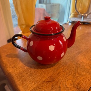 May include: A red enamel teapot with white polka dots. The teapot has a black handle and spout, and a matching lid. The teapot is sitting on a wooden surface.