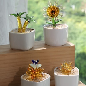May include: Four small white ceramic pots each hold a unique glass plant sculpture. The sculptures include palm trees, a sunflower, and other abstract floral designs. The glass plants are yellow, green, and blue, and the pots are filled with small white stones.