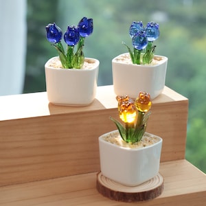 May include: Three small white square ceramic pots each hold glass tulip flowers. The tulips are blue and amber, with green stems and leaves. The pots sit on a wooden surface.