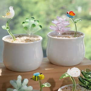 Pode incluir: Duas pequenas jarras de cerâmica branca com plantas decorativas de vidro. Uma jarra apresenta um pássaro de vidro, a outra uma flor rosa com uma borboleta. As jarras são preenchidas com pequenas pedras, criando uma cena de jardim em miniatura.