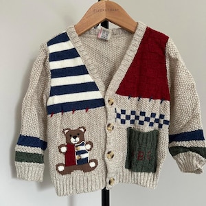 May include: Beige knit cardigan with a teddy bear applique and color block design. Features navy and white stripes, a red panel, and a green pocket with "ABC" embroidery. Button-down front with wooden buttons.