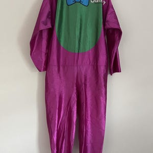 Könnte beinhalten: Ein violetter Langarm-Jumpsuit mit einem großen grünen ovalen Einsatz auf der Brust. Das Kleidungsstück zeigt eine blaue Fliege als Grafik und den in Weiß gedruckten Schriftzug "Barney" im oberen Brustbereich. Der Stoff hat eine glatte, glänzende Oberfläche.