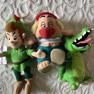 May include: Three plush toys featuring a character in a green tunic and hat, a character with a red hat and blue and white striped shirt, and a green crocodile with an open mouth showing white teeth and a pink tongue, all made of soft fabric.
