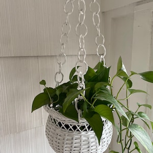 May include: A white wicker hanging planter with a chain, filled with green leafy plants. The planter is round and has a woven design. The chain is made of large, white, interlocking links. The plant has long, trailing vines.
