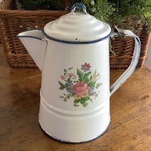 May include: White enamelware coffee pot with a floral design featuring pink roses and blue accents. The pot has a lid, handle, and spout, with blue trim. A vintage kitchenware item.