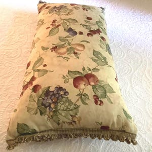 Puede incluir: Cojín decorativo rectangular con fondo crema y estampado botánico de frutas, bayas y hojas en tonos rojos, verdes y morados. El cojín tiene un ribete con flecos en el borde inferior.