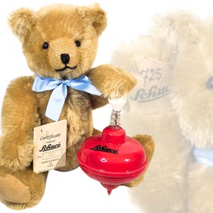 Op de afbeelding: Een bruin teddybeer met een lichtblauw lint om zijn nek houdt een rode en zilveren tol vast. De beer heeft een label met de tekst "Schuco" en "Certificate". Op de tol staat het woord "Schuco".