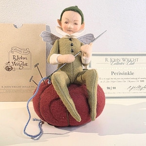 May include: A collectible R. John Wright doll named Periwinkle, seated on a red pincushion, holding a needle and thread. The doll is dressed in green and beige clothing with small wings. The doll is accompanied by a certificate of authenticity and a cardboard box.