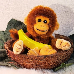 Op de afbeelding: Een bruine pluchen aap met een gele banaan zit in een bruine kokosnootschaal. De aap heeft een lachend gezicht en zwarte ogen. De kokosnootschaal ligt op een groen blad.