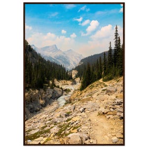 May include: A framed landscape print of a mountain valley with a river. The sky is blue with white clouds. The valley is filled with green trees and a rocky path. The print is in a brown frame.