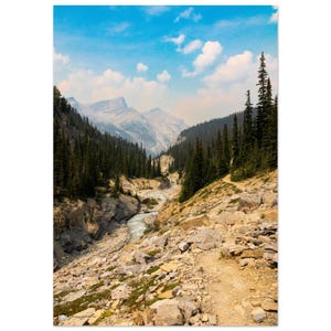 May include: A scenic landscape print featuring a mountain range, a river, and a path winding through a valley. The sky is blue with scattered clouds. The valley is filled with evergreen trees and rocky terrain.