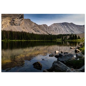Pode incluir: Fotografia de paisagem de um lago de montanha. A água clara reflete as montanhas, árvores e céu circundantes. Grandes rochas são visíveis em primeiro plano. O céu é azul com algumas nuvens. As montanhas são cinzentas e castanhas.