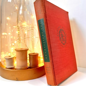 May include: A vintage book titled "The Ambassadors" with a red cover and green spine. The book is propped up next to a decorative glass cloche with string lights and wooden spools on a gold base.
