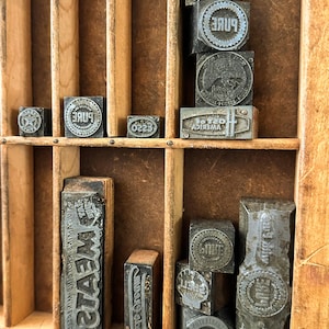 May include: A collection of vintage metal letterpress printing blocks arranged in a wooden partitioned tray. The blocks feature various embossed logos and text including Shell, Pure, Esso, and Meats, with some stacked vertically in the compartments.