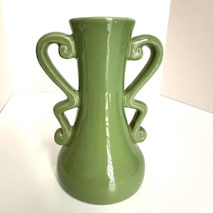May include: A green ceramic vase with two ornate handles. The vase has a bulbous base and a flared top. The handles are curved and decorative, adding a unique touch to the design. The vase is a solid, glossy green color.