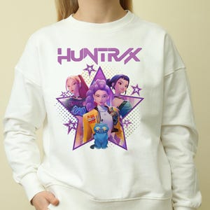 May include: White sweatshirt featuring a graphic of three stylized women and a blue cat inside a purple star. The word "HUNTRAX" is printed in purple above the star. The design includes stars and a dotted background.