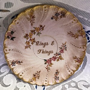 May include: A vintage, cream-coloured ceramic ring dish with a gold rim and floral accents. The dish features the text "Rings & Things" in a gold font, surrounded by delicate flower and leaf patterns. The dish has a scalloped edge.