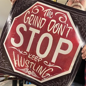 May include: A red and white sign with the text "The Grind Don't Stop" and "Keep Hustling". The sign is in a stop sign shape with a dark brown border and spiderweb design. The sign has a vintage, hand-drawn style.