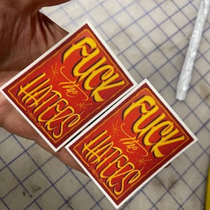 May include: Two rectangular stickers featuring a red background with a yellow and black border. The text "FUCK the HATERS" is printed in a stylized yellow font with black outlines, accompanied by small star accents on the red surface.